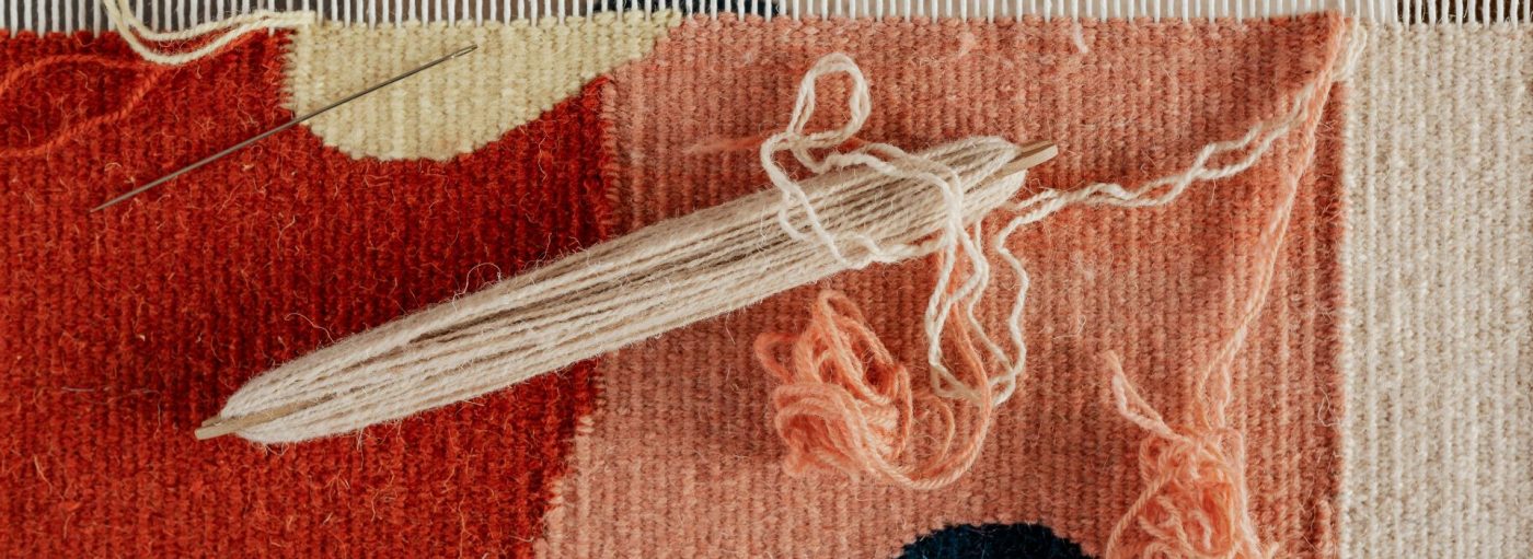 A weaving loom part way through a wool project that is red, yellow, peach and beige.